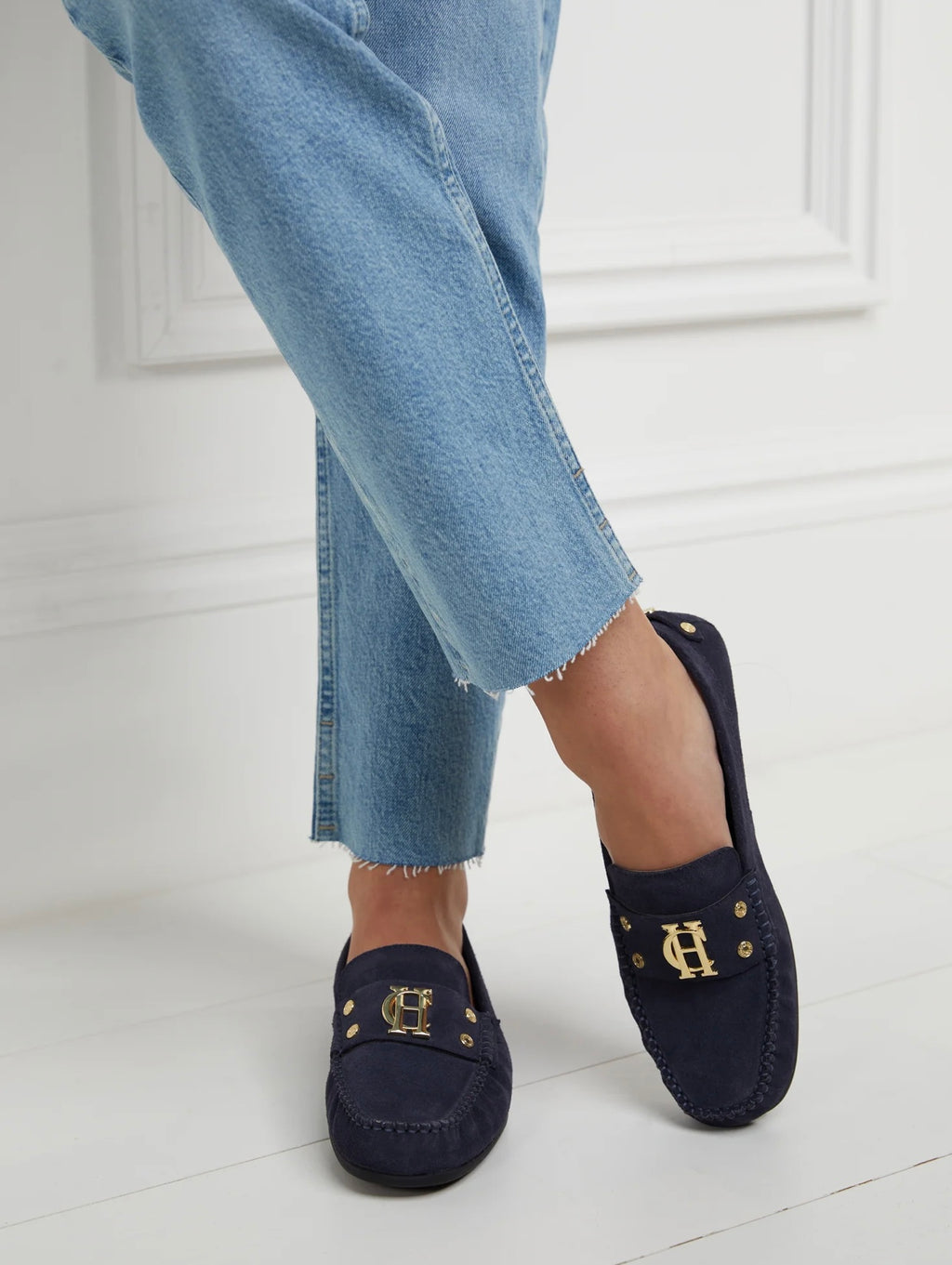 Navy loafers with gold accents worn with light blue jeans on a white floor.