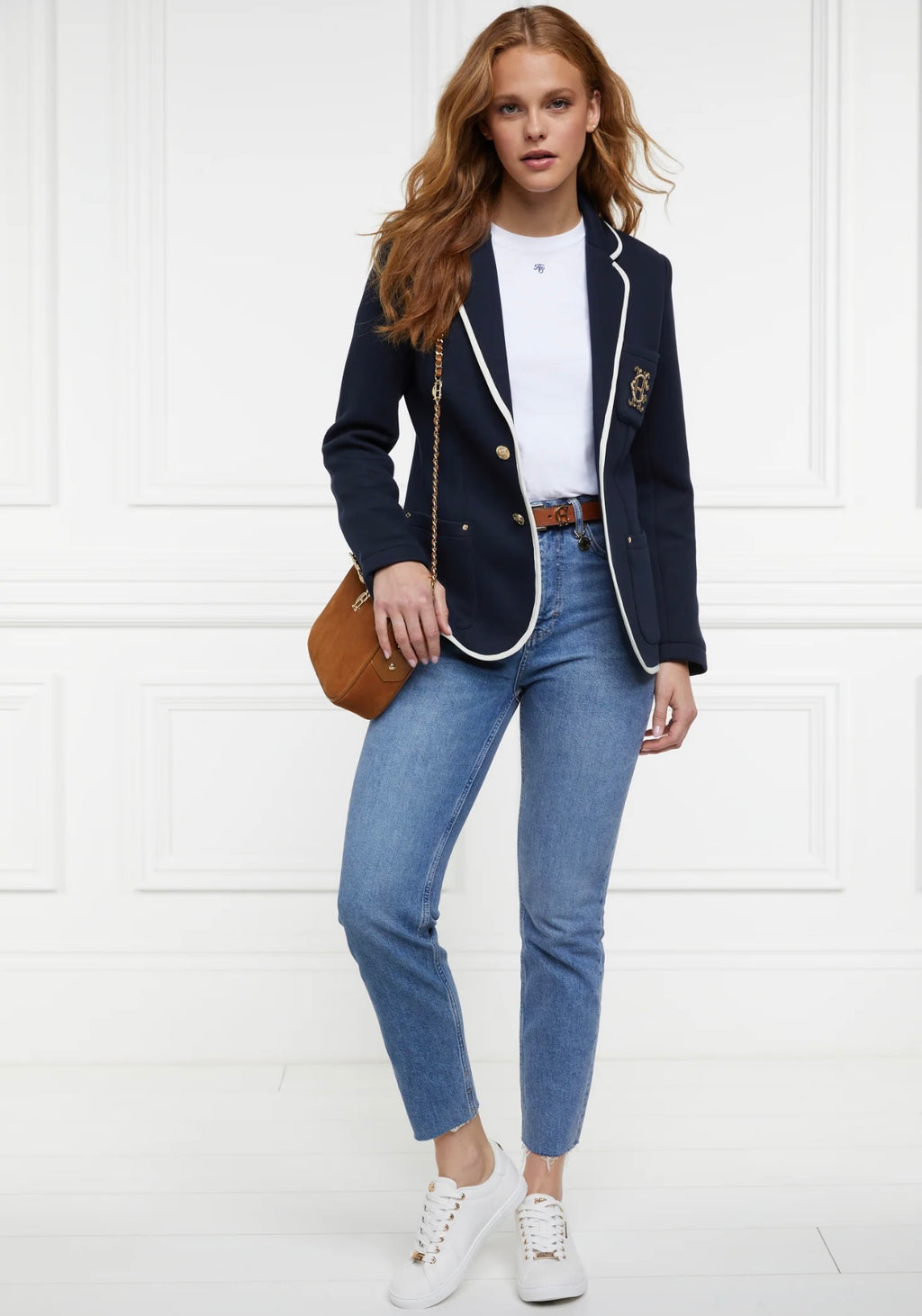 Woman wearing a navy blazer, white shirt, blue jeans, and white sneakers on a white background