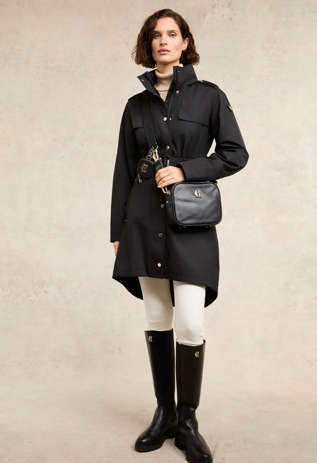 Woman wearing a black coat, white leggings, and black rain boots holding a black bag against a beige background