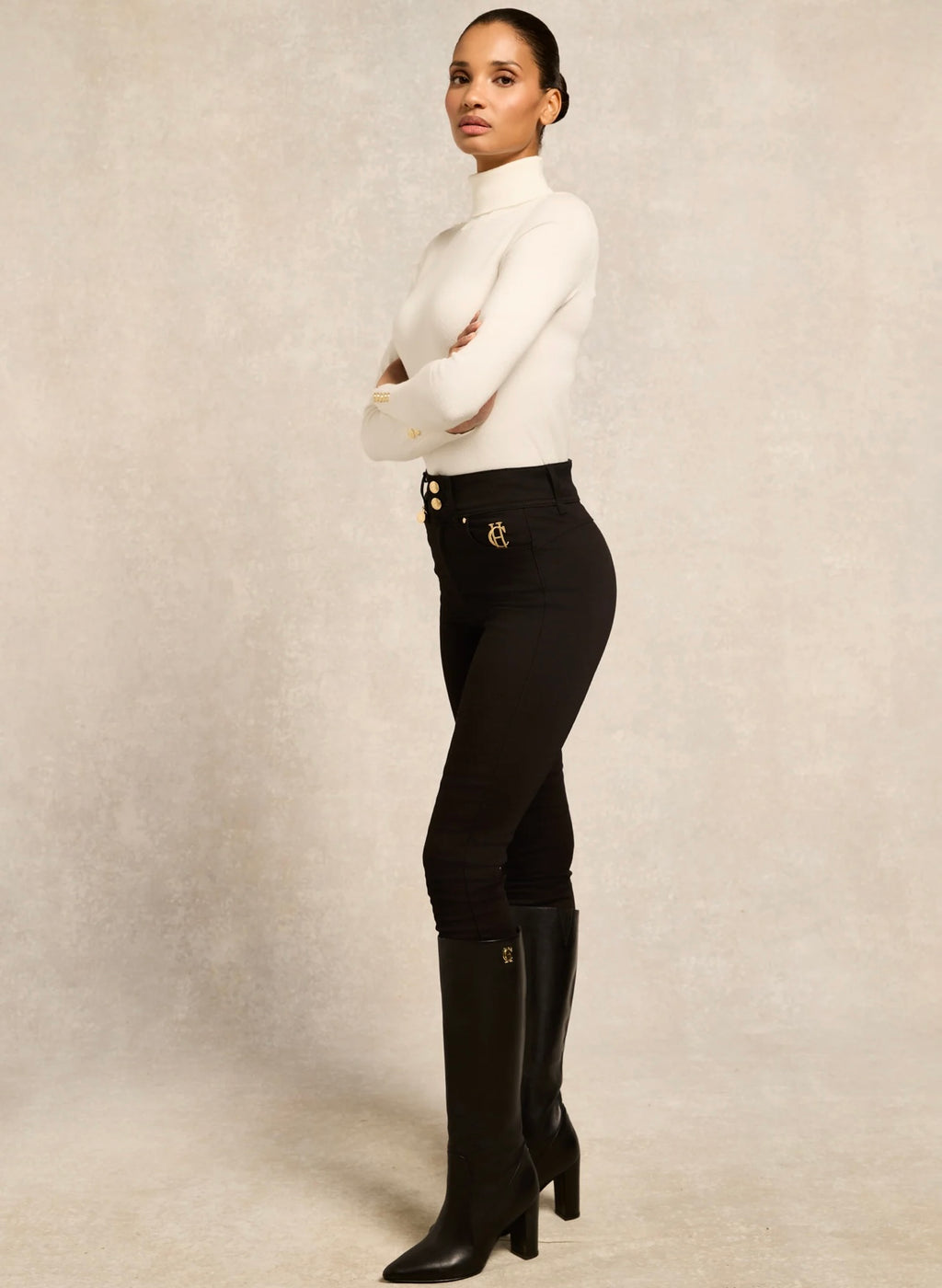 Person wearing a white turtleneck and black riding pants with knee-high boots on a neutral background