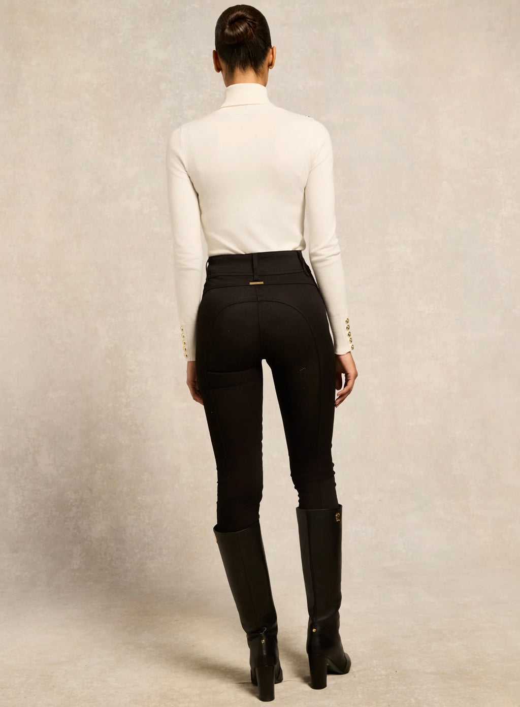 Person wearing black riding pants and boots with a white shirt on a neutral background