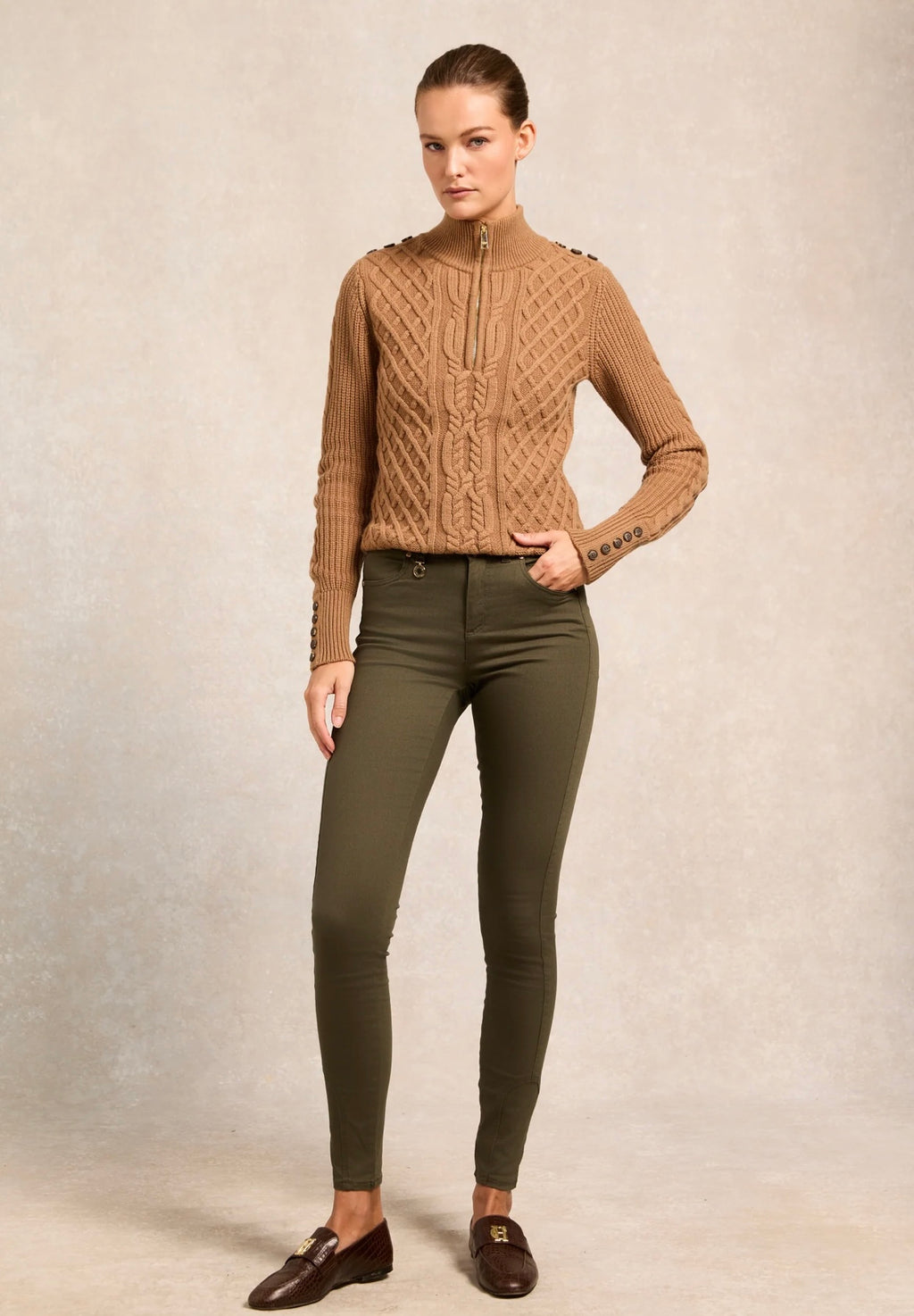 Woman wearing a brown cable knit sweater and green pants against a beige background