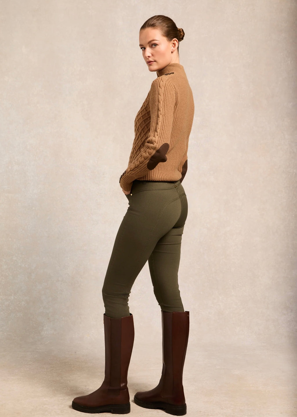 Woman wearing a brown knitted sweater, green riding pants, and brown riding boots on a neutral background