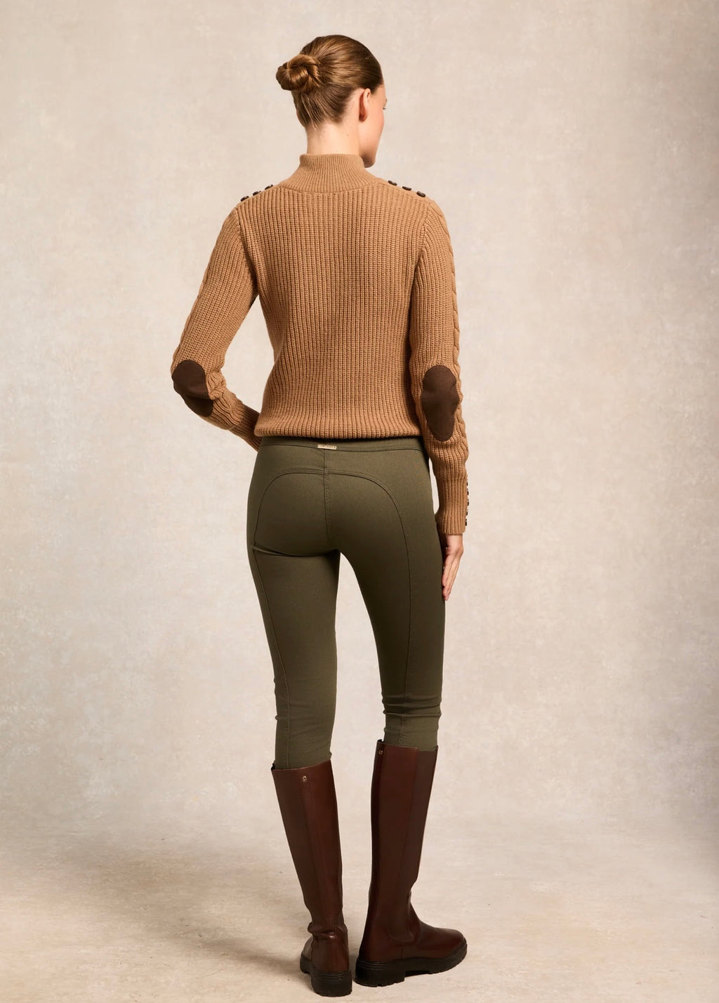 Person wearing a brown ribbed sweater, green riding pants, and brown riding boots against a beige background