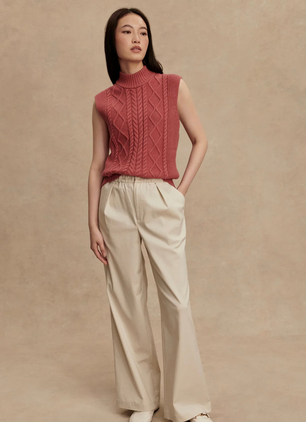 Woman wearing a pink knitted sleeveless sweater and beige pants against a beige background