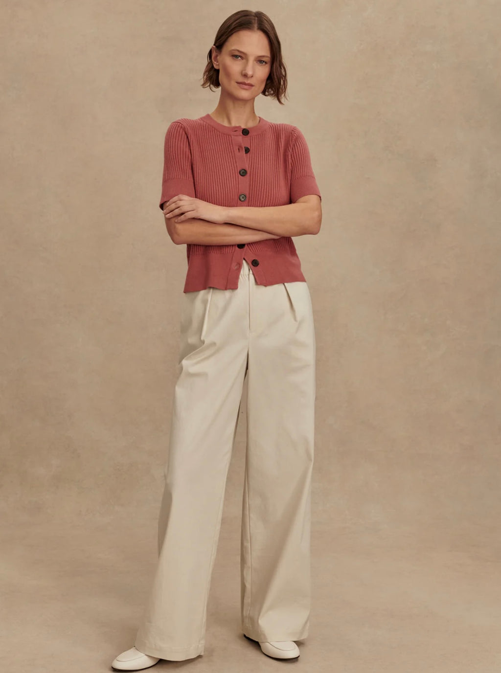 Woman wearing a pink cropped cardigan and white pants against a beige background