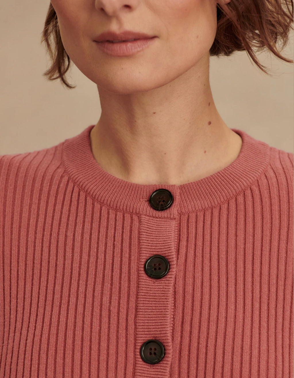 Close-up of a person wearing a pink ribbed sweater with black buttons on a beige background