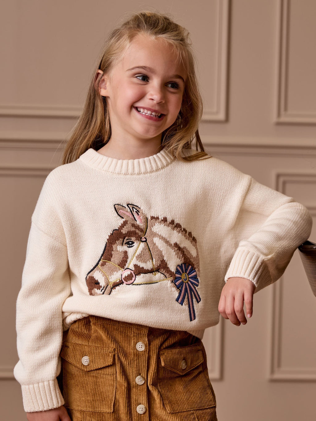 Young girl wearing a cream sweater with a donkey design against a beige wall.