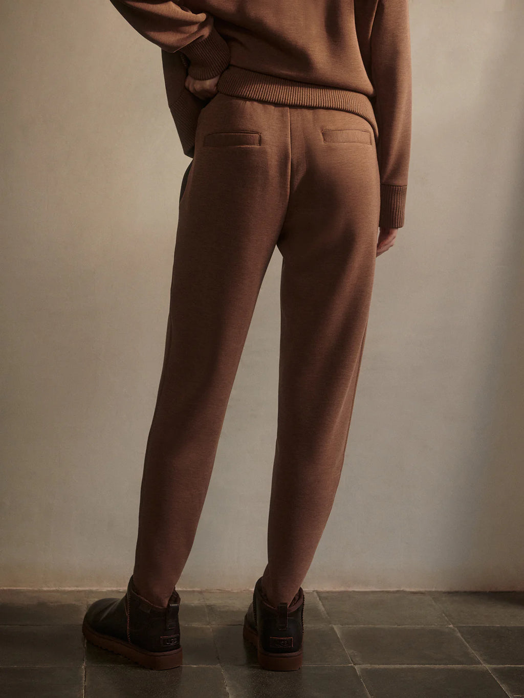Person wearing brown pants and sweater against a plain wall.