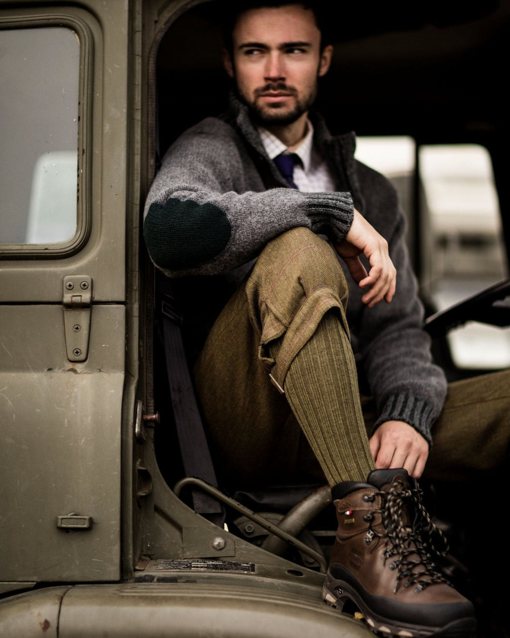 Man sitting inside a vintage vehicle wearing a gray sweater, brown pants, and hiking boots.