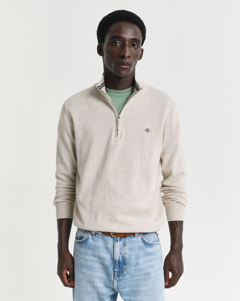 Man wearing a beige sweater and light blue jeans against a white background
