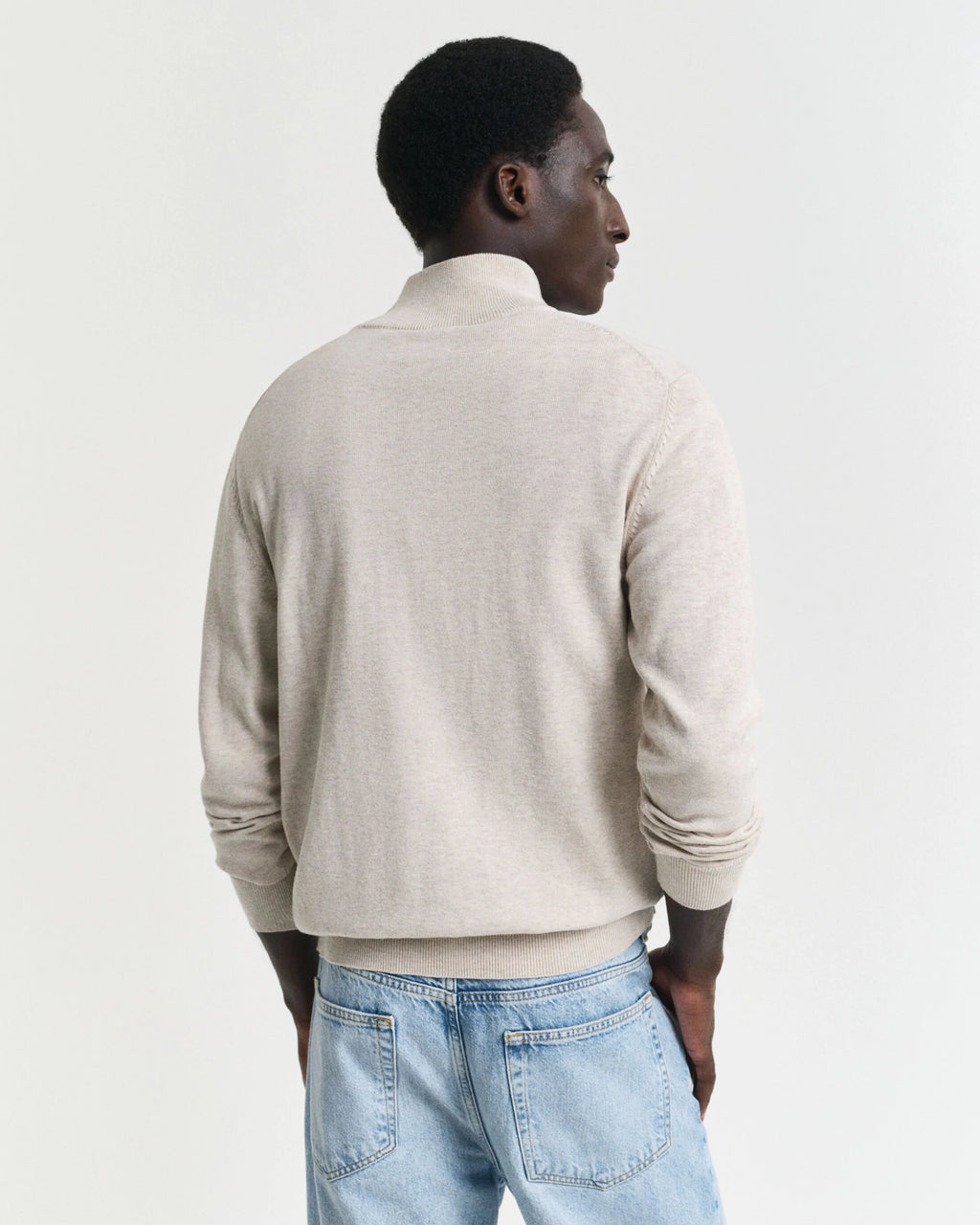Man wearing a beige sweater and light blue jeans on a plain background