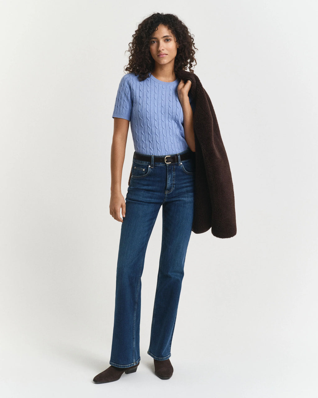 Woman wearing a blue top and jeans holding a brown coat on a white background